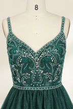 A photograph of a mannequin showcasing a floor-length, sleeveless, dark green evening gown. The gown features a V-neckline embellished with intricate beadwork and a fitted bodice that transitions into a voluminous tulle skirt. The background is plain and off-white, emphasizing the gown's elegant design.9133b.2