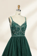 A photograph of a mannequin showcasing a floor-length, sleeveless, dark green evening gown. The gown features a V-neckline embellished with intricate beadwork and a fitted bodice that transitions into a voluminous tulle skirt. The background is plain and off-white, emphasizing the gown's elegant design.9133b.3