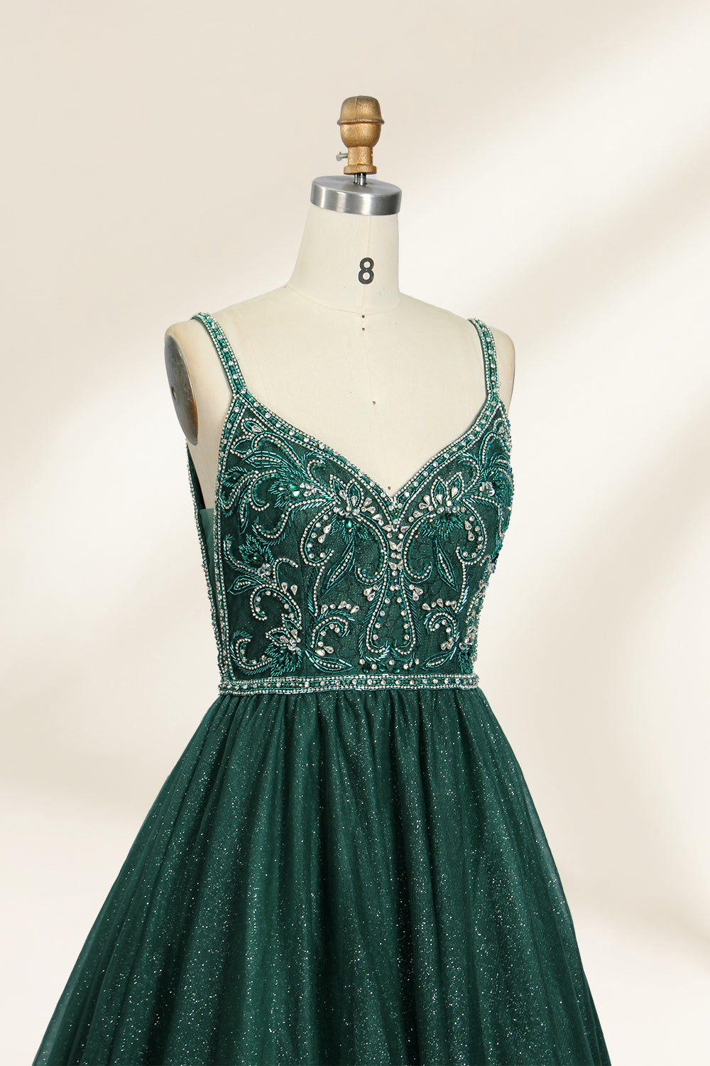 A photograph of a mannequin showcasing a floor-length, sleeveless, dark green evening gown. The gown features a V-neckline embellished with intricate beadwork and a fitted bodice that transitions into a voluminous tulle skirt. The background is plain and off-white, emphasizing the gown's elegant design.9133b.3
