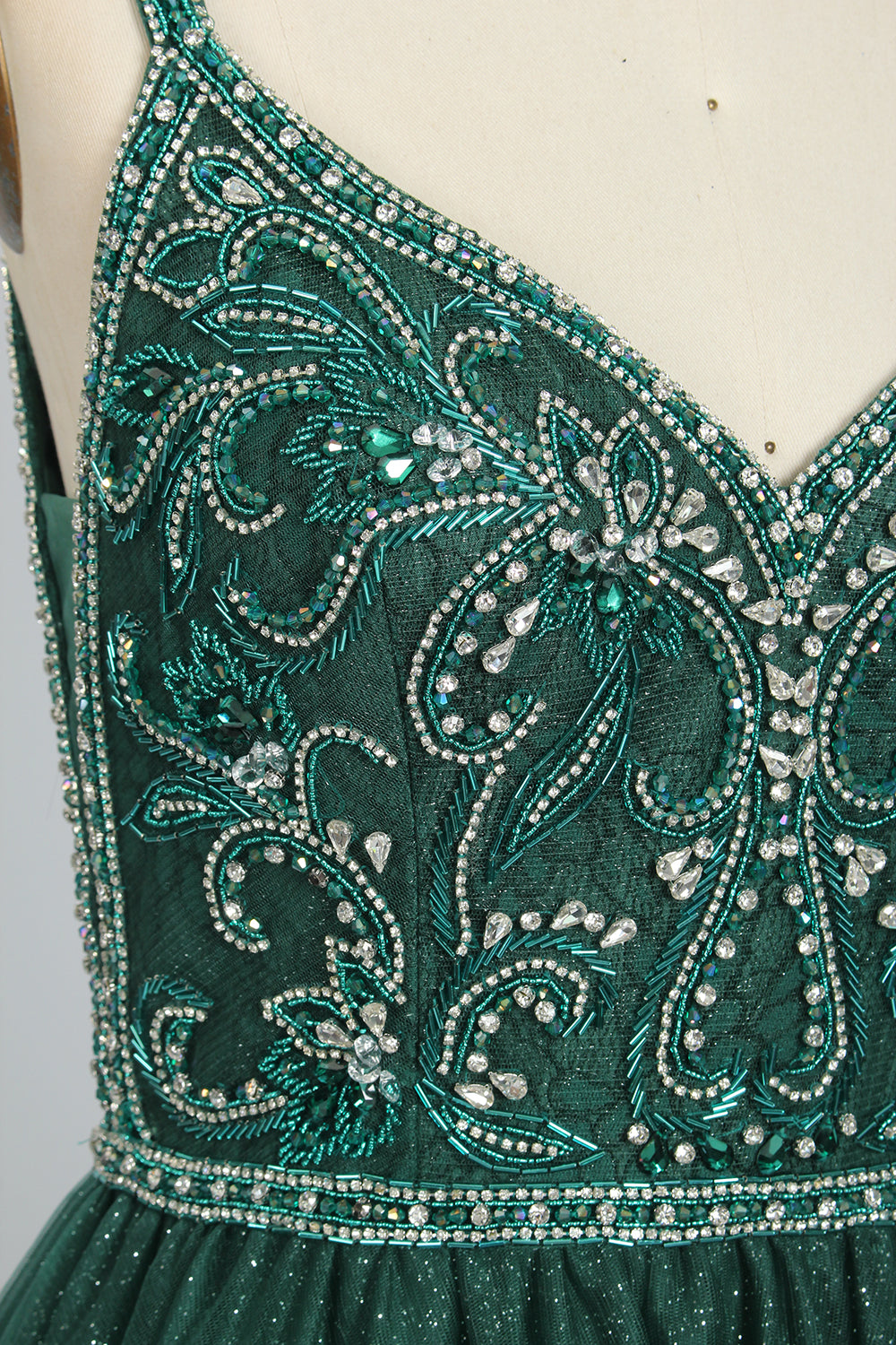 A photograph of a mannequin showcasing a floor-length, sleeveless, dark green evening gown. The gown features a V-neckline embellished with intricate beadwork and a fitted bodice that transitions into a voluminous tulle skirt. The background is plain and off-white, emphasizing the gown's elegant design.9133b.4