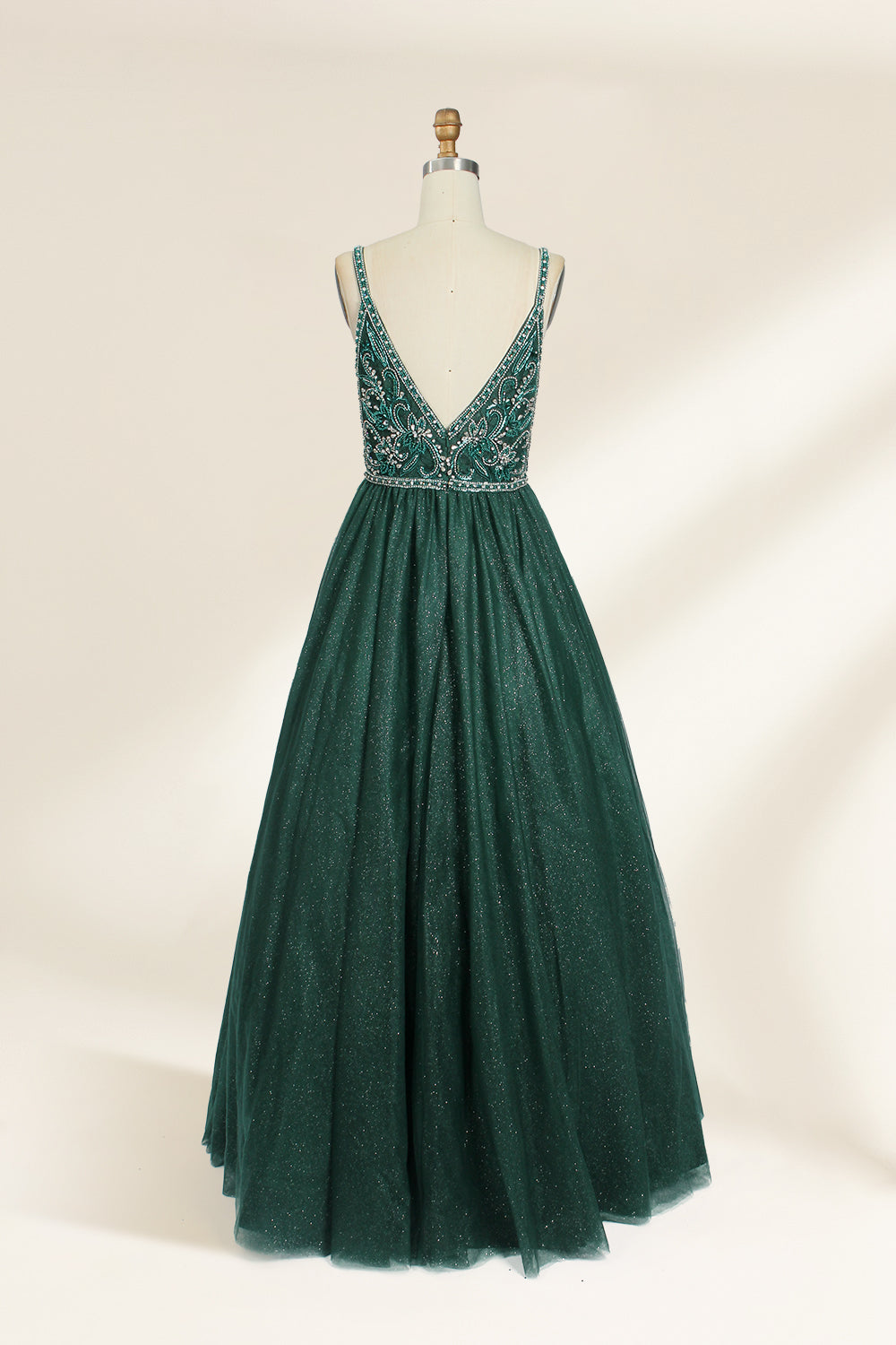 A photograph of a mannequin showcasing a floor-length, sleeveless, dark green evening gown. The gown features a V-neckline embellished with intricate beadwork and a fitted bodice that transitions into a voluminous tulle skirt. The background is plain and off-white, emphasizing the gown's elegant design.9133b.5