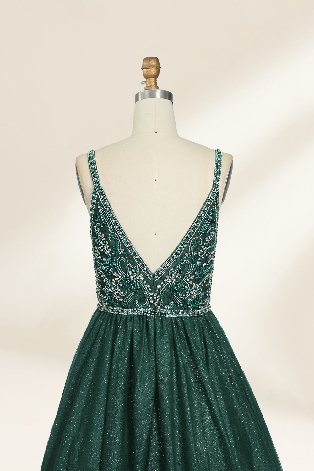 A photograph of a mannequin showcasing a floor-length, sleeveless, dark green evening gown. The gown features a V-neckline embellished with intricate beadwork and a fitted bodice that transitions into a voluminous tulle skirt. The background is plain and off-white, emphasizing the gown's elegant design.9133b.6