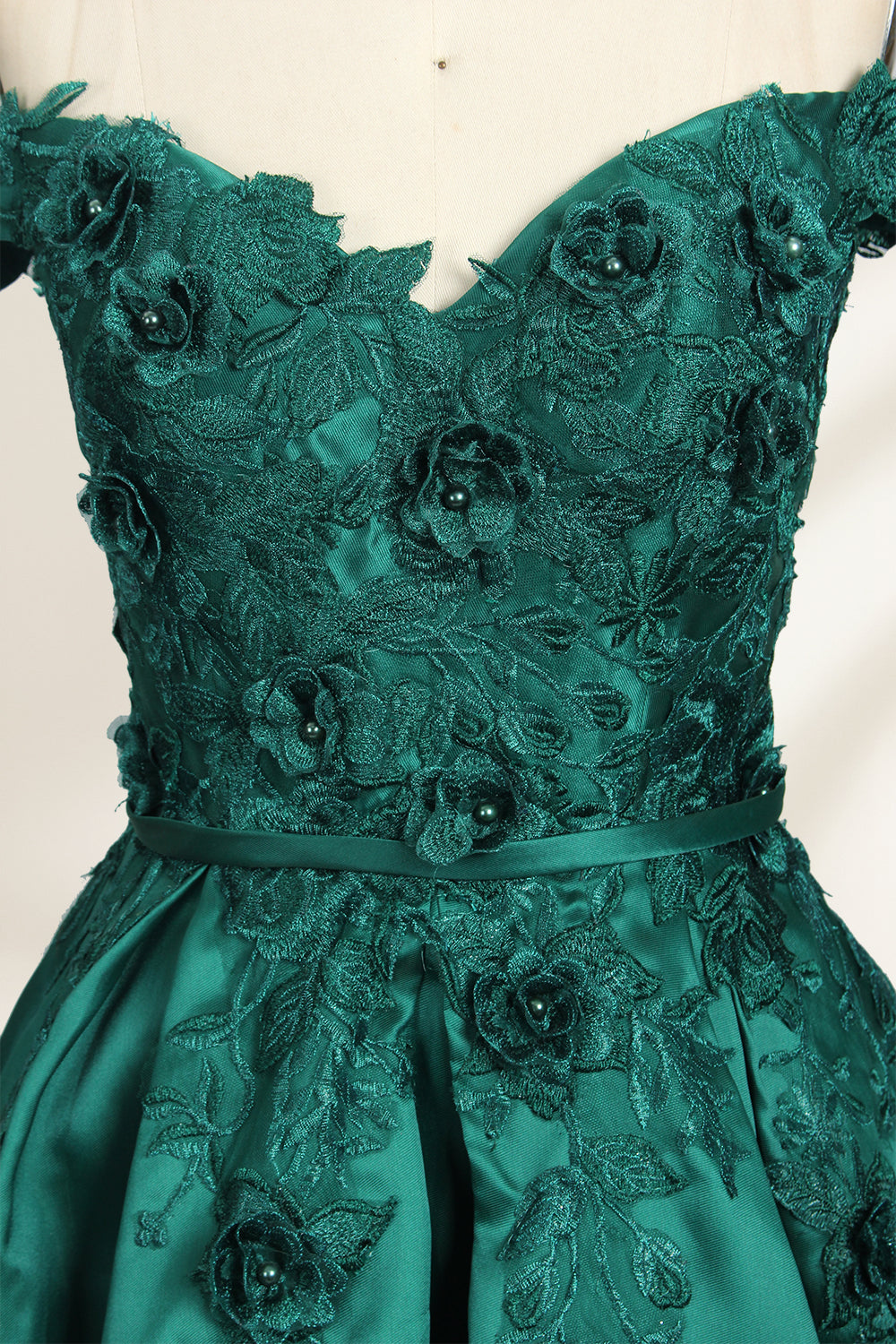 This is a photograph of a stunning, off-the-shoulder, floor-length evening gown displayed on a dress form. The gown is a rich, emerald green satin with intricate black floral lace embroidery on the bodice and hem. The dress features a sweetheart neckline, accentuating the bust, and a fitted waist that flares into a full, flowing skirt. The dress form is beige, with a wooden head and neck, and a metal neck ring. The background is a plain, light cream color, ensuring the dress remains the focal point.06
