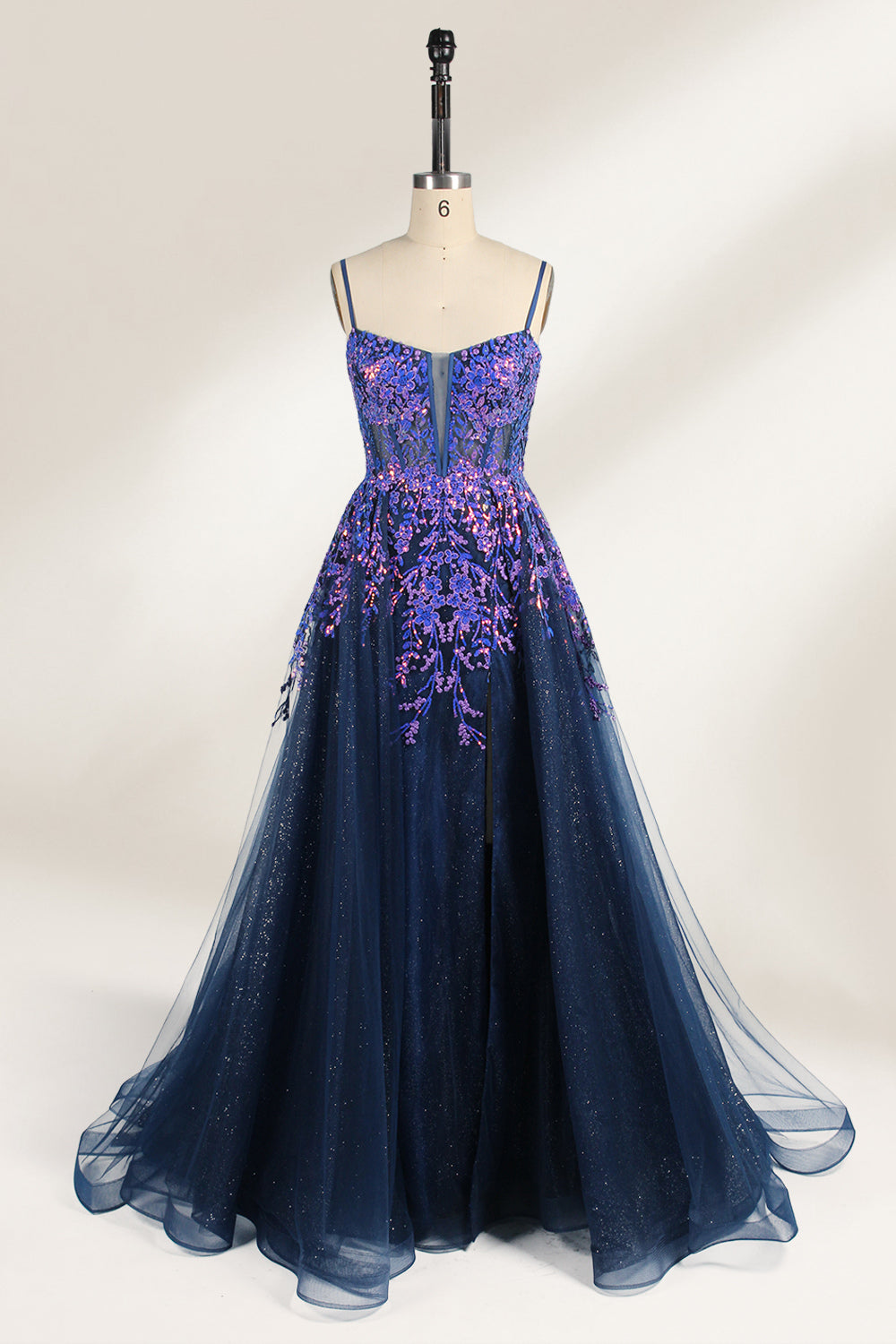 A photograph of a dark navy blue, floor-length evening gown displayed on a dress form. The gown features intricate floral embroidery in shades of purple and pink on the bodice, transitioning to a sheer, sparkling tulle skirt. The bodice is fitted with spaghetti straps, and the dress form is positioned against a plain, off-white background, highlighting the gown's elegant and luxurious design.1