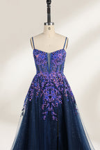 A photograph of a dark navy blue, floor-length evening gown displayed on a dress form. The gown features intricate floral embroidery in shades of purple and pink on the bodice, transitioning to a sheer, sparkling tulle skirt. The bodice is fitted with spaghetti straps, and the dress form is positioned against a plain, off-white background, highlighting the gown's elegant and luxurious design.5