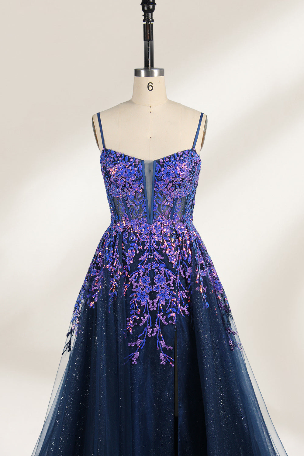 A photograph of a dark navy blue, floor-length evening gown displayed on a dress form. The gown features intricate floral embroidery in shades of purple and pink on the bodice, transitioning to a sheer, sparkling tulle skirt. The bodice is fitted with spaghetti straps, and the dress form is positioned against a plain, off-white background, highlighting the gown's elegant and luxurious design.5