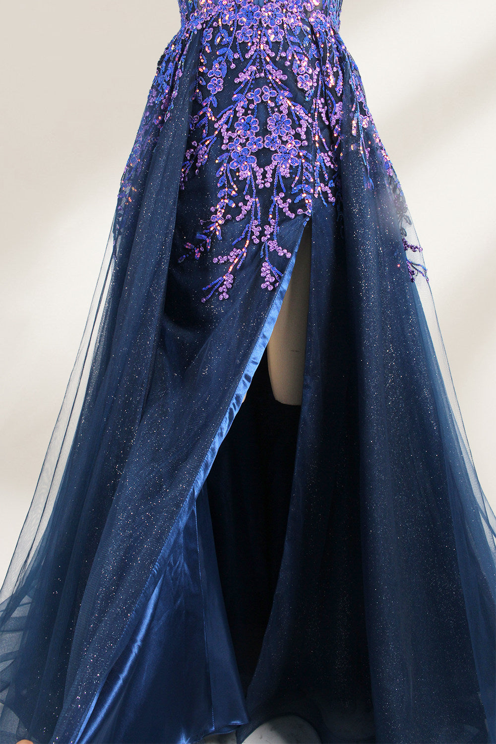 A photograph of a dark navy blue, floor-length evening gown displayed on a dress form. The gown features intricate floral embroidery in shades of purple and pink on the bodice, transitioning to a sheer, sparkling tulle skirt. The bodice is fitted with spaghetti straps, and the dress form is positioned against a plain, off-white background, highlighting the gown's elegant and luxurious design.4