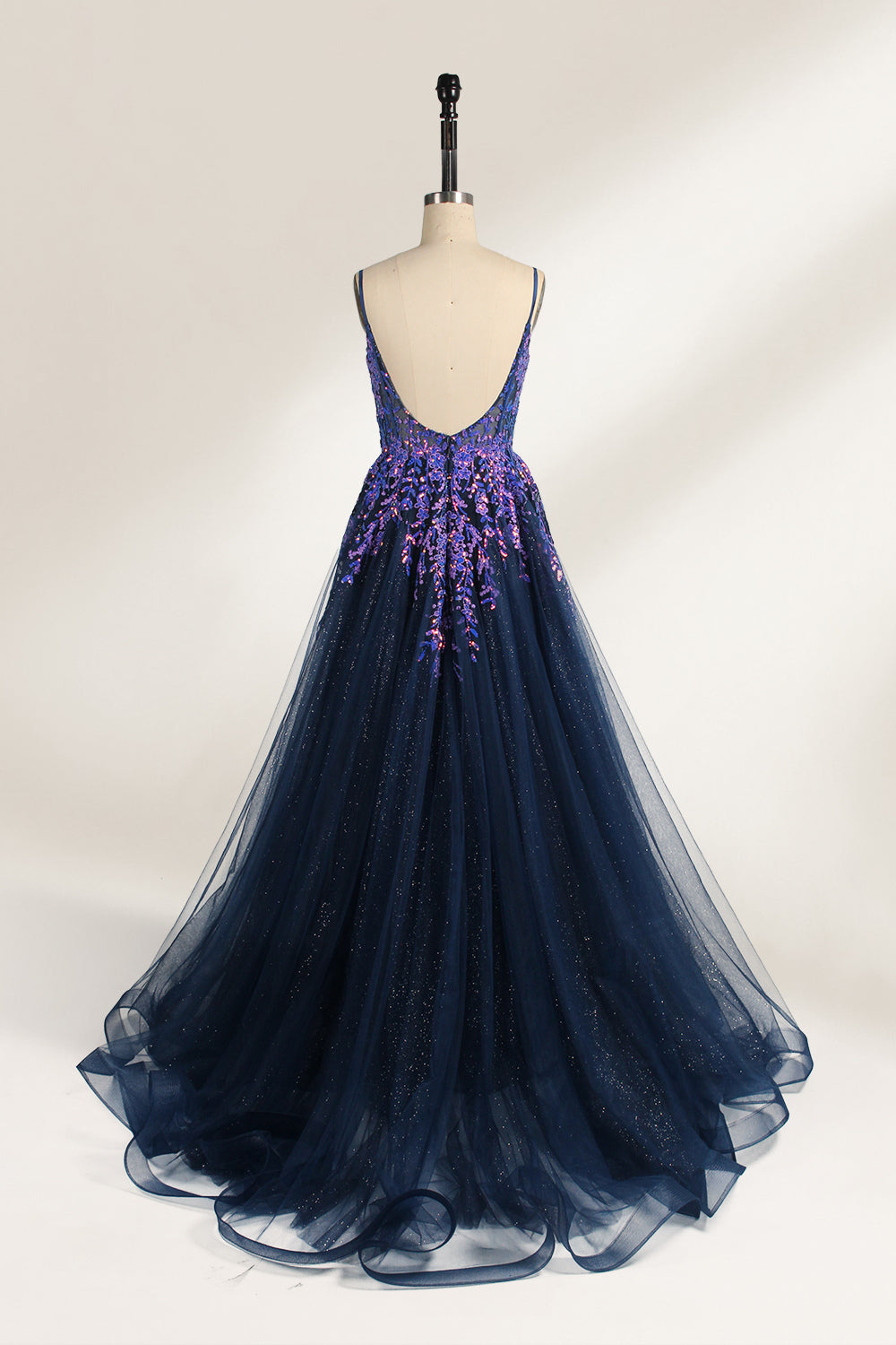 A photograph of a dark navy blue, floor-length evening gown displayed on a dress form. The gown features intricate floral embroidery in shades of purple and pink on the bodice, transitioning to a sheer, sparkling tulle skirt. The bodice is fitted with spaghetti straps, and the dress form is positioned against a plain, off-white background, highlighting the gown's elegant and luxurious design.2