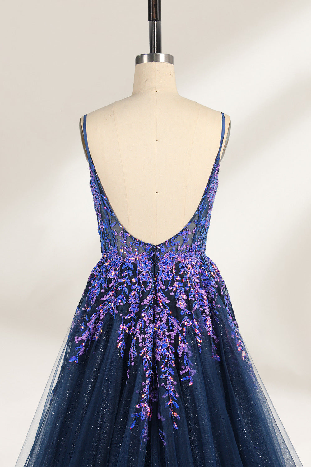 A photograph of a dark navy blue, floor-length evening gown displayed on a dress form. The gown features intricate floral embroidery in shades of purple and pink on the bodice, transitioning to a sheer, sparkling tulle skirt. The bodice is fitted with spaghetti straps, and the dress form is positioned against a plain, off-white background, highlighting the gown's elegant and luxurious design.6