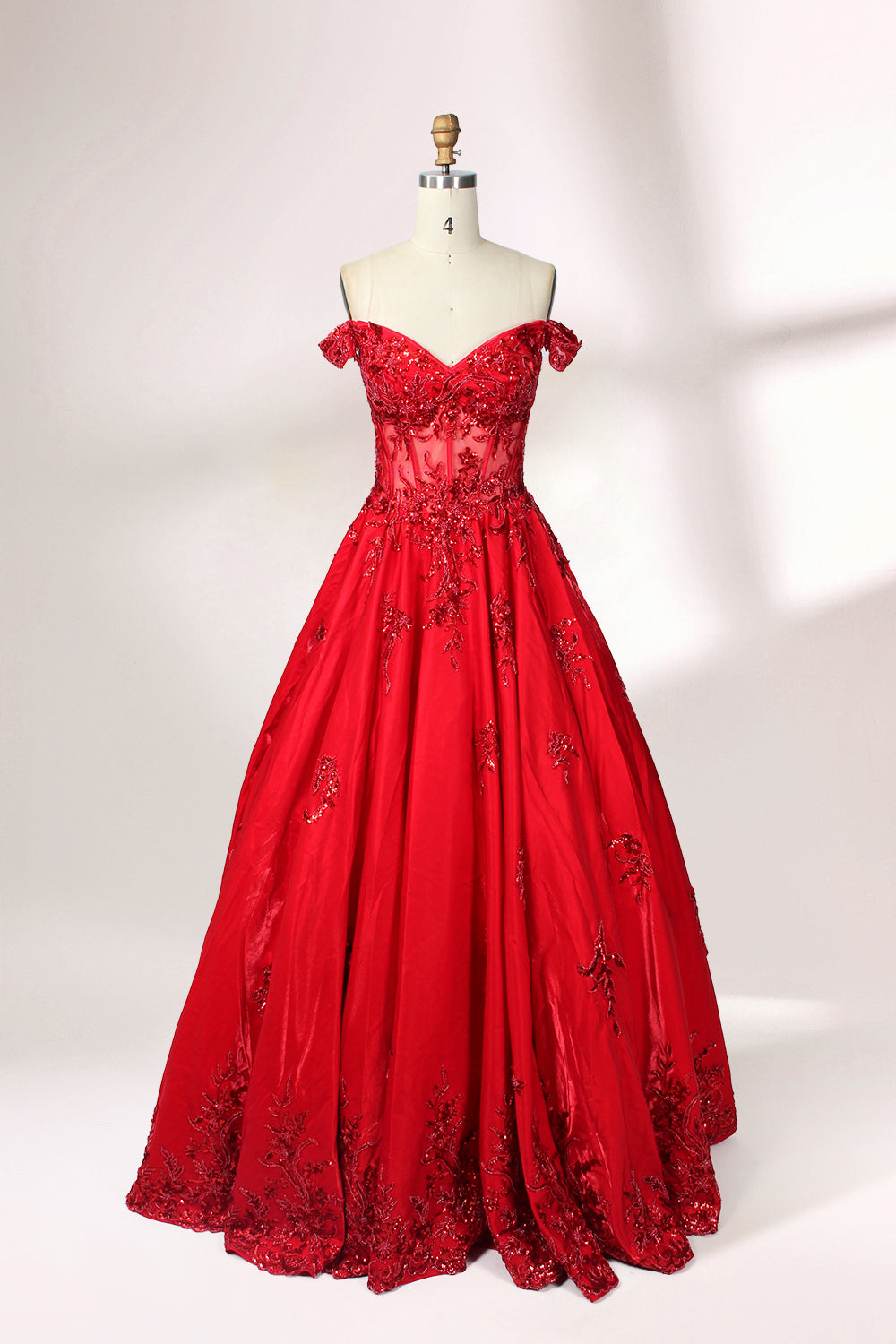 Photograph of an elegant, off-the-shoulder red ball gown on a white dress form. The dress features intricate, sparkling beadwork on the bodice and a voluminous, flowing skirt adorned with delicate floral patterns. The background is a plain white studio setup with soft shadows, emphasizing the dress's vibrant color and luxurious texture.01