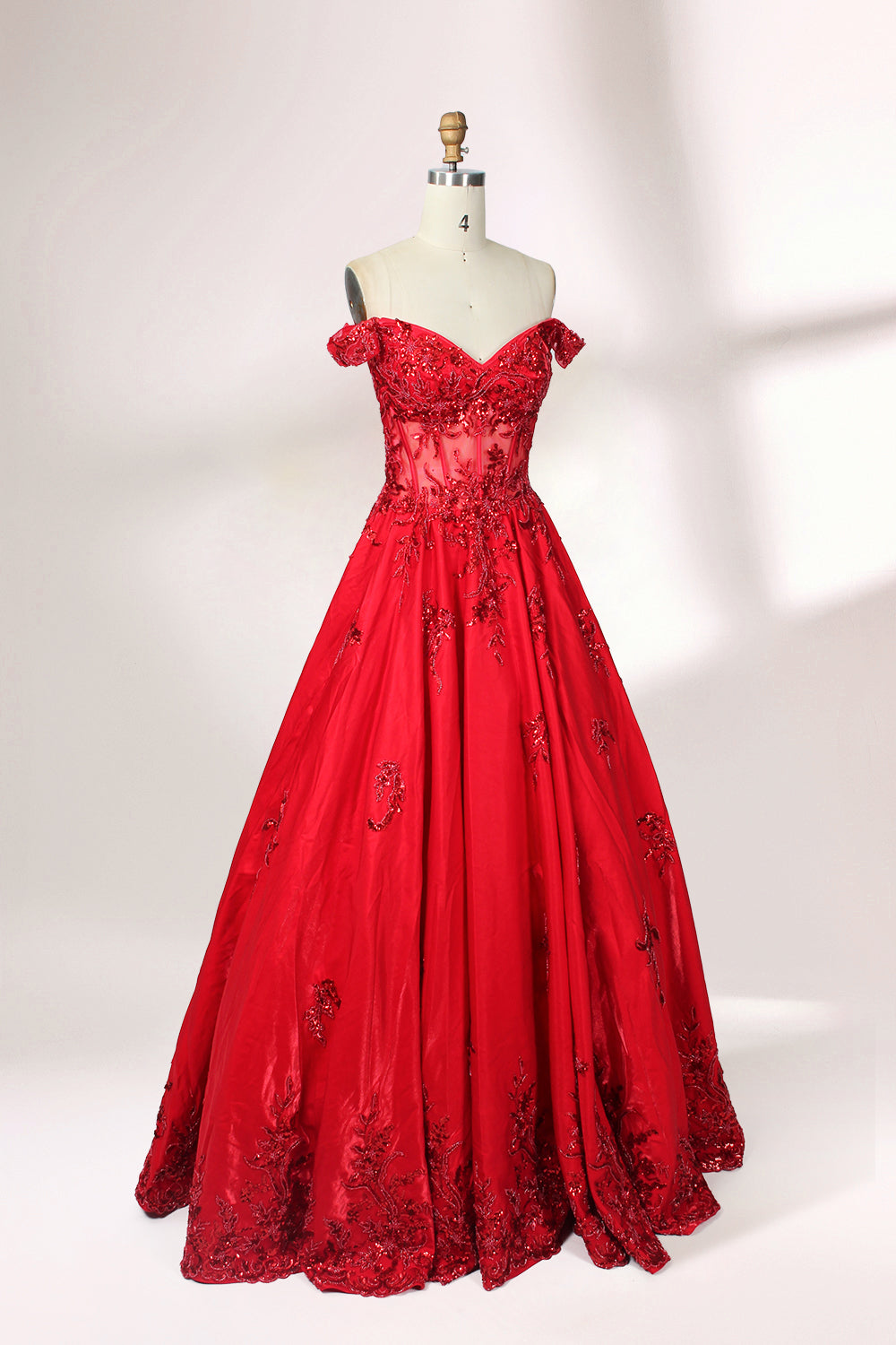 Photograph of an elegant, off-the-shoulder red ball gown on a white dress form. The dress features intricate, sparkling beadwork on the bodice and a voluminous, flowing skirt adorned with delicate floral patterns. The background is a plain white studio setup with soft shadows, emphasizing the dress's vibrant color and luxurious texture.04