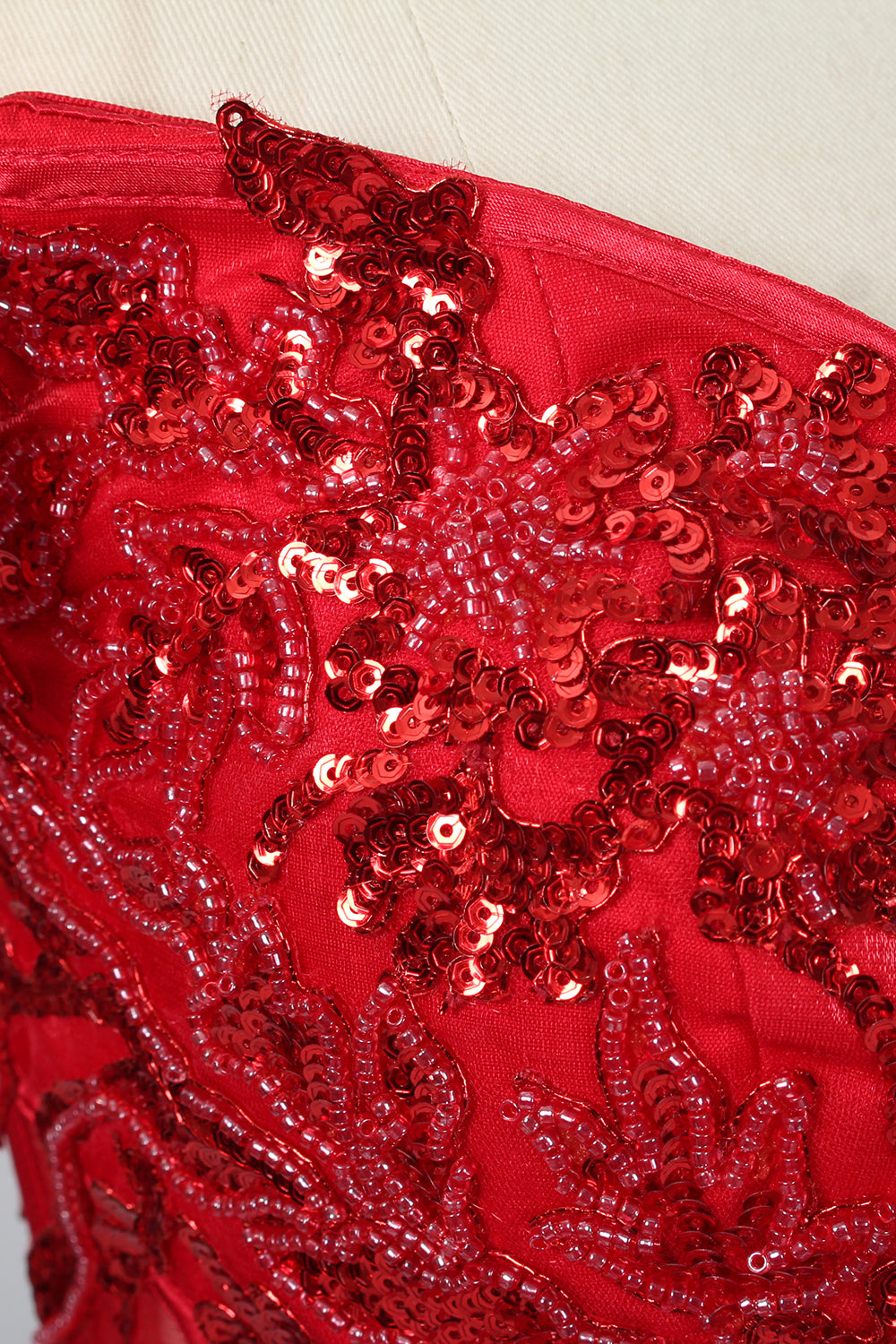 Photograph of an elegant, off-the-shoulder red ball gown on a white dress form. The dress features intricate, sparkling beadwork on the bodice and a voluminous, flowing skirt adorned with delicate floral patterns. The background is a plain white studio setup with soft shadows, emphasizing the dress's vibrant color and luxurious texture.06