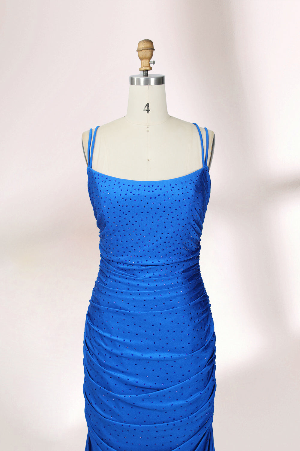 Photograph of a blue, floor-length evening gown displayed on a white dress form. The gown is sleeveless with a fitted bodice and a flared skirt, featuring thin shoulder straps and a textured, ruched design. The dress form is placed against a plain, off-white background, casting soft shadows.04