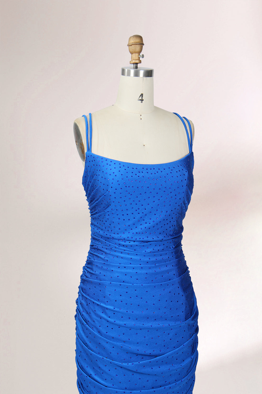 Photograph of a blue, floor-length evening gown displayed on a white dress form. The gown is sleeveless with a fitted bodice and a flared skirt, featuring thin shoulder straps and a textured, ruched design. The dress form is placed against a plain, off-white background, casting soft shadows.05