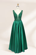 Photograph of a mannequin wearing a floor-length, deep V-neck emerald green satin gown with intricate lace detailing around the neckline and bodice. The gown features a fitted bodice and a flowing, pleated skirt. The mannequin stands on a wooden base against a plain, light beige background, highlighting the gown's elegant and luxurious design.01