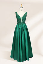 Photograph of a mannequin wearing a floor-length, deep V-neck emerald green satin gown with intricate lace detailing around the neckline and bodice. The gown features a fitted bodice and a flowing, pleated skirt. The mannequin stands on a wooden base against a plain, light beige background, highlighting the gown's elegant and luxurious design.01