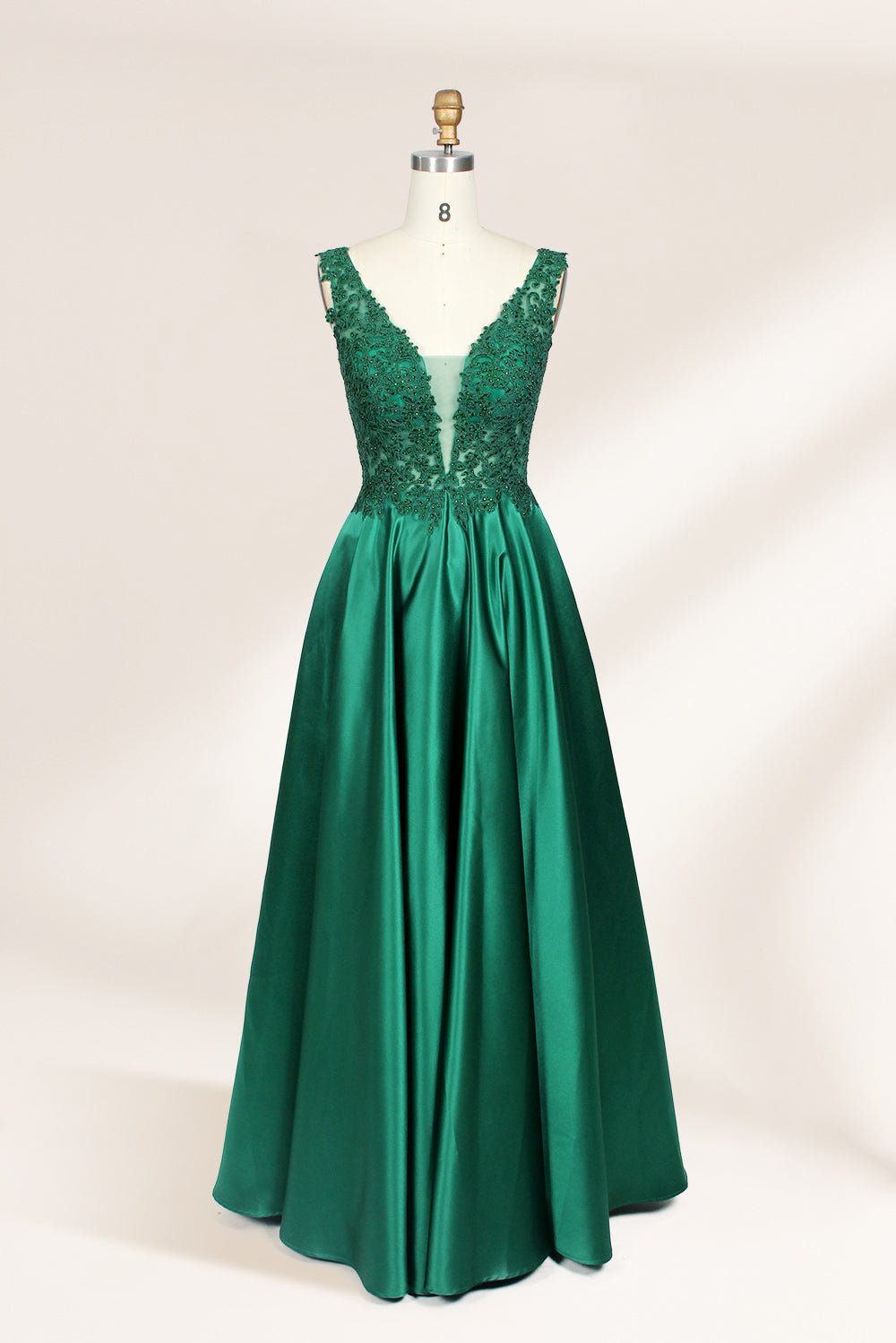 Photograph of a mannequin wearing a floor-length, deep V-neck emerald green satin gown with intricate lace detailing around the neckline and bodice. The gown features a fitted bodice and a flowing, pleated skirt. The mannequin stands on a wooden base against a plain, light beige background, highlighting the gown's elegant and luxurious design.01