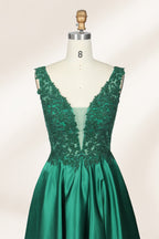 Photograph of a mannequin wearing a floor-length, deep V-neck emerald green satin gown with intricate lace detailing around the neckline and bodice. The gown features a fitted bodice and a flowing, pleated skirt. The mannequin stands on a wooden base against a plain, light beige background, highlighting the gown's elegant and luxurious design.03