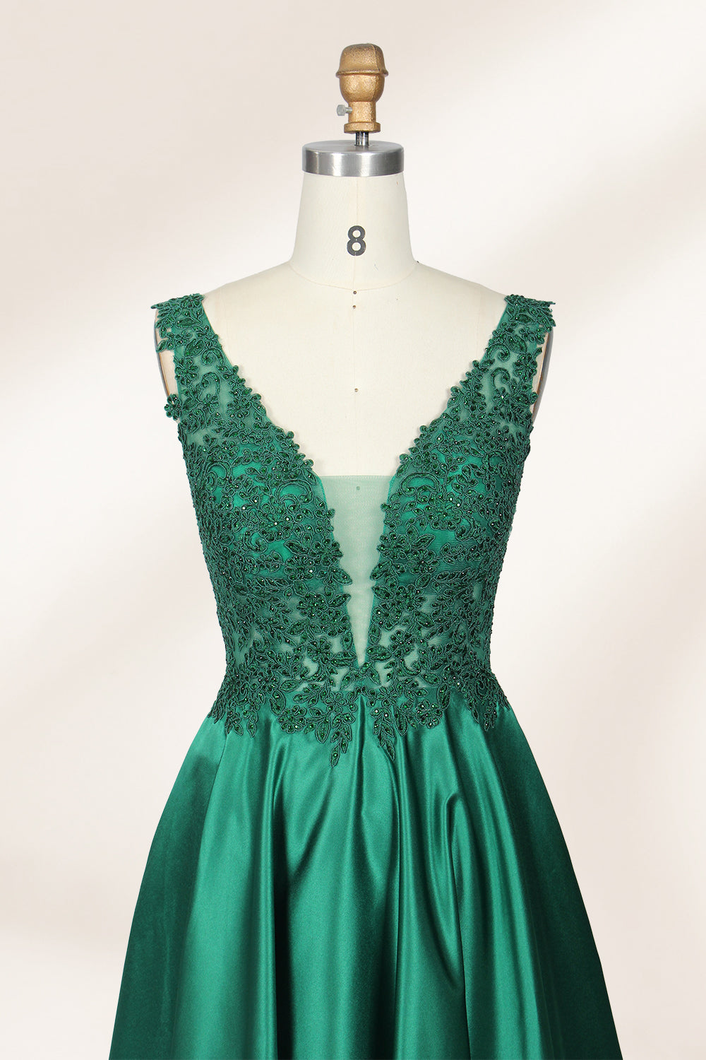 Photograph of a mannequin wearing a floor-length, deep V-neck emerald green satin gown with intricate lace detailing around the neckline and bodice. The gown features a fitted bodice and a flowing, pleated skirt. The mannequin stands on a wooden base against a plain, light beige background, highlighting the gown's elegant and luxurious design.03