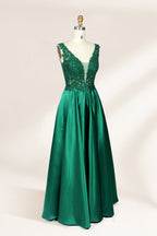 Photograph of a mannequin wearing a floor-length, deep V-neck emerald green satin gown with intricate lace detailing around the neckline and bodice. The gown features a fitted bodice and a flowing, pleated skirt. The mannequin stands on a wooden base against a plain, light beige background, highlighting the gown's elegant and luxurious design.04