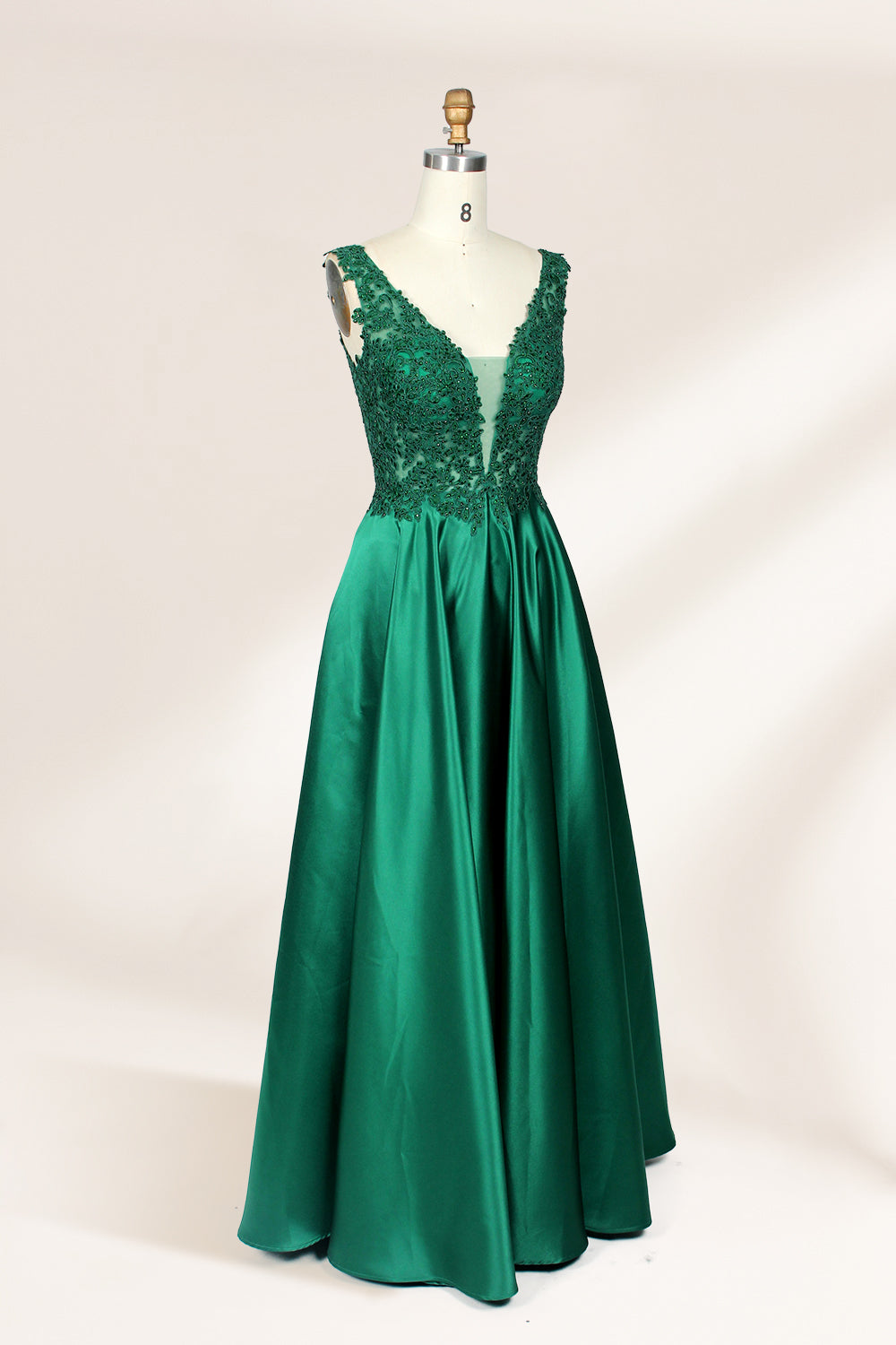 Photograph of a mannequin wearing a floor-length, deep V-neck emerald green satin gown with intricate lace detailing around the neckline and bodice. The gown features a fitted bodice and a flowing, pleated skirt. The mannequin stands on a wooden base against a plain, light beige background, highlighting the gown's elegant and luxurious design.04