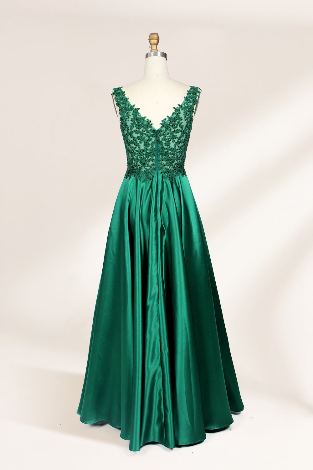 Photograph of a mannequin wearing a floor-length, deep V-neck emerald green satin gown with intricate lace detailing around the neckline and bodice. The gown features a fitted bodice and a flowing, pleated skirt. The mannequin stands on a wooden base against a plain, light beige background, highlighting the gown's elegant and luxurious design.02
