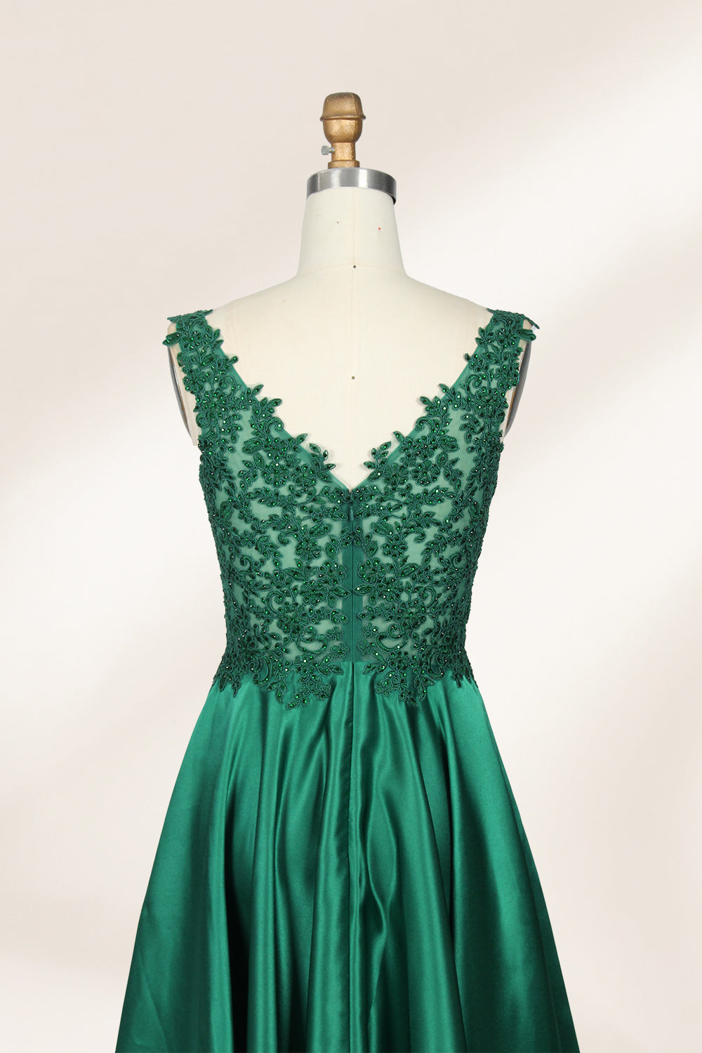 Photograph of a mannequin wearing a floor-length, deep V-neck emerald green satin gown with intricate lace detailing around the neckline and bodice. The gown features a fitted bodice and a flowing, pleated skirt. The mannequin stands on a wooden base against a plain, light beige background, highlighting the gown's elegant and luxurious design.05