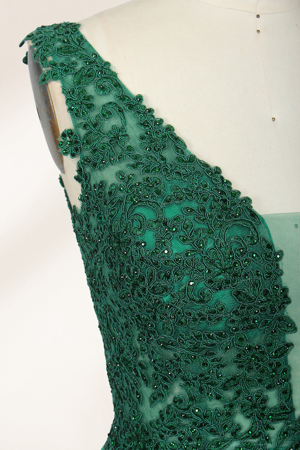 Photograph of a mannequin wearing a floor-length, deep V-neck emerald green satin gown with intricate lace detailing around the neckline and bodice. The gown features a fitted bodice and a flowing, pleated skirt. The mannequin stands on a wooden base against a plain, light beige background, highlighting the gown's elegant and luxurious design.06