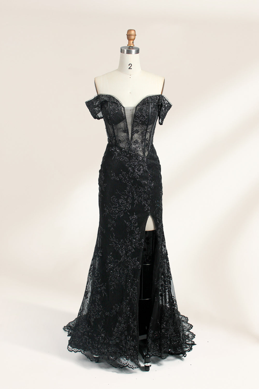 This photograph features a mannequin wearing an elegant, floor-length black lace gown. The gown has a sweetheart neckline, off-the-shoulder sleeves, and a high slit on one side, revealing a black thigh-high stocking. The intricate lace pattern is delicate and ornate, adding texture and visual interest. The mannequin stands against a plain, light beige background, emphasizing the dress's details. The number "2" is displayed on the mannequin's chest.01