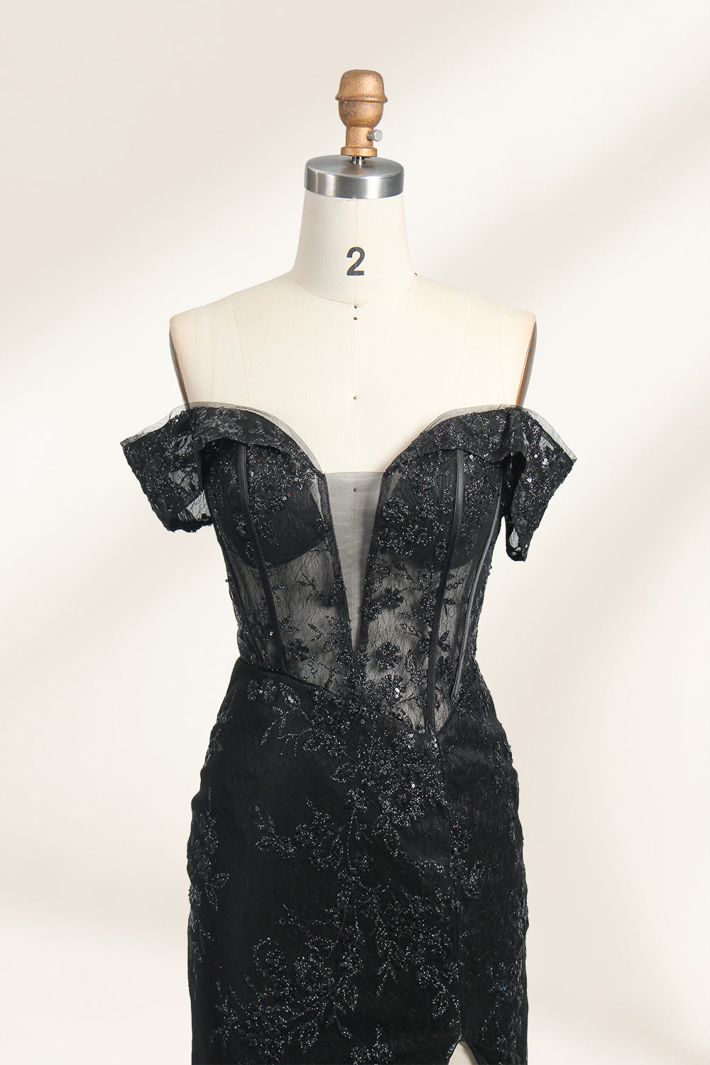This photograph features a mannequin wearing an elegant, floor-length black lace gown. The gown has a sweetheart neckline, off-the-shoulder sleeves, and a high slit on one side, revealing a black thigh-high stocking. The intricate lace pattern is delicate and ornate, adding texture and visual interest. The mannequin stands against a plain, light beige background, emphasizing the dress's details. The number "2" is displayed on the mannequin's chest.03
