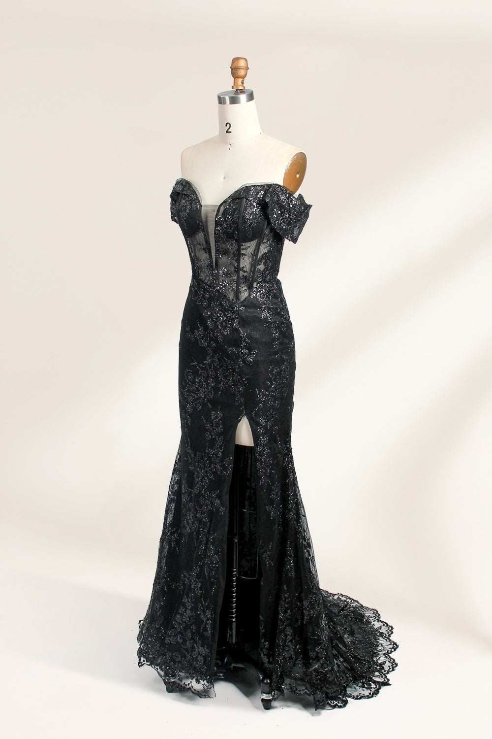 This photograph features a mannequin wearing an elegant, floor-length black lace gown. The gown has a sweetheart neckline, off-the-shoulder sleeves, and a high slit on one side, revealing a black thigh-high stocking. The intricate lace pattern is delicate and ornate, adding texture and visual interest. The mannequin stands against a plain, light beige background, emphasizing the dress's details. The number "2" is displayed on the mannequin's chest.05