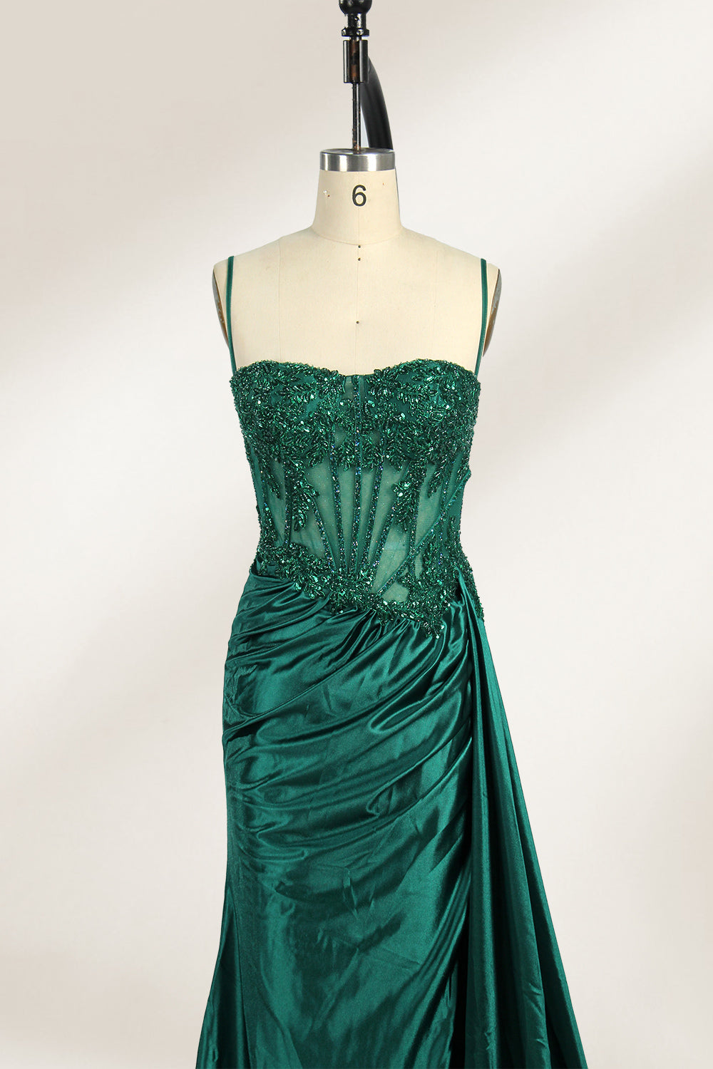 Photograph of an elegant, strapless green satin evening gown on a dress form. The dress features a fitted bodice adorned with intricate beading and lacework, transitioning into a flowing, ruched skirt that cascades to the floor. The gown's deep emerald green color exudes luxury and sophistication. The background is a plain, off-white studio setting, highlighting the dress's opulence. The dress form is positioned centrally, with the dress's front view prominently displayed.5
