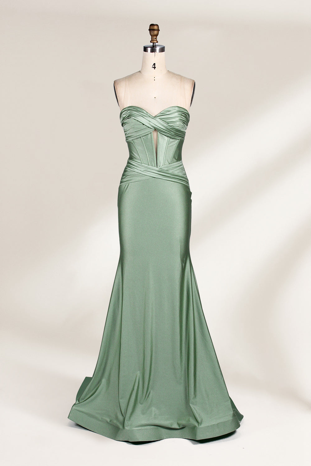 This photograph showcases a floor-length, strapless gown in a soft, mint green hue. The dress features a fitted bodice with a sweetheart neckline and a delicate, crisscrossed fabric pattern, accentuating the bust. The skirt flares out elegantly, ending in a subtle train. The gown is displayed on a mannequin with a wooden head and neck, set against a plain, light beige background. The fabric appears to be satin or silk, giving it a smooth, lustrous texture. 1