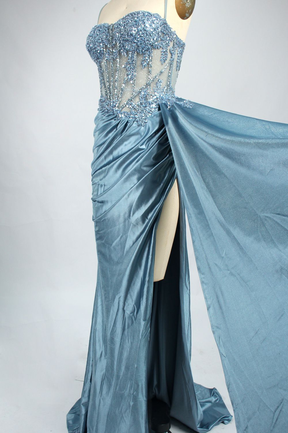 Photograph of an elegant, strapless green satin evening gown on a dress form. The dress features a fitted bodice adorned with intricate beading and lacework, transitioning into a flowing, ruched skirt that cascades to the floor. The gown's deep emerald green color exudes luxury and sophistication. The background is a plain, off-white studio setting, highlighting the dress's opulence. The dress form is positioned centrally, with the dress's front view prominently displayed.8