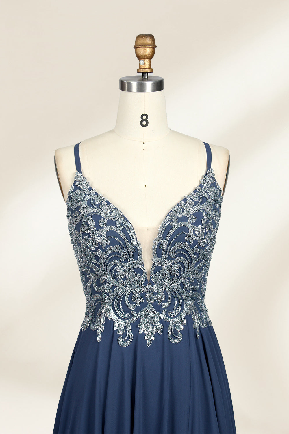 Photograph of a mannequin showcasing an elegant, floor-length evening gown. The gown features a deep V-neckline adorned with intricate blue floral embroidery, transitioning into a flowing, dark blue chiffon skirt. The mannequin stands against a plain, cream-colored backdrop. The dress exudes sophistication and luxury, perfect for formal events.03