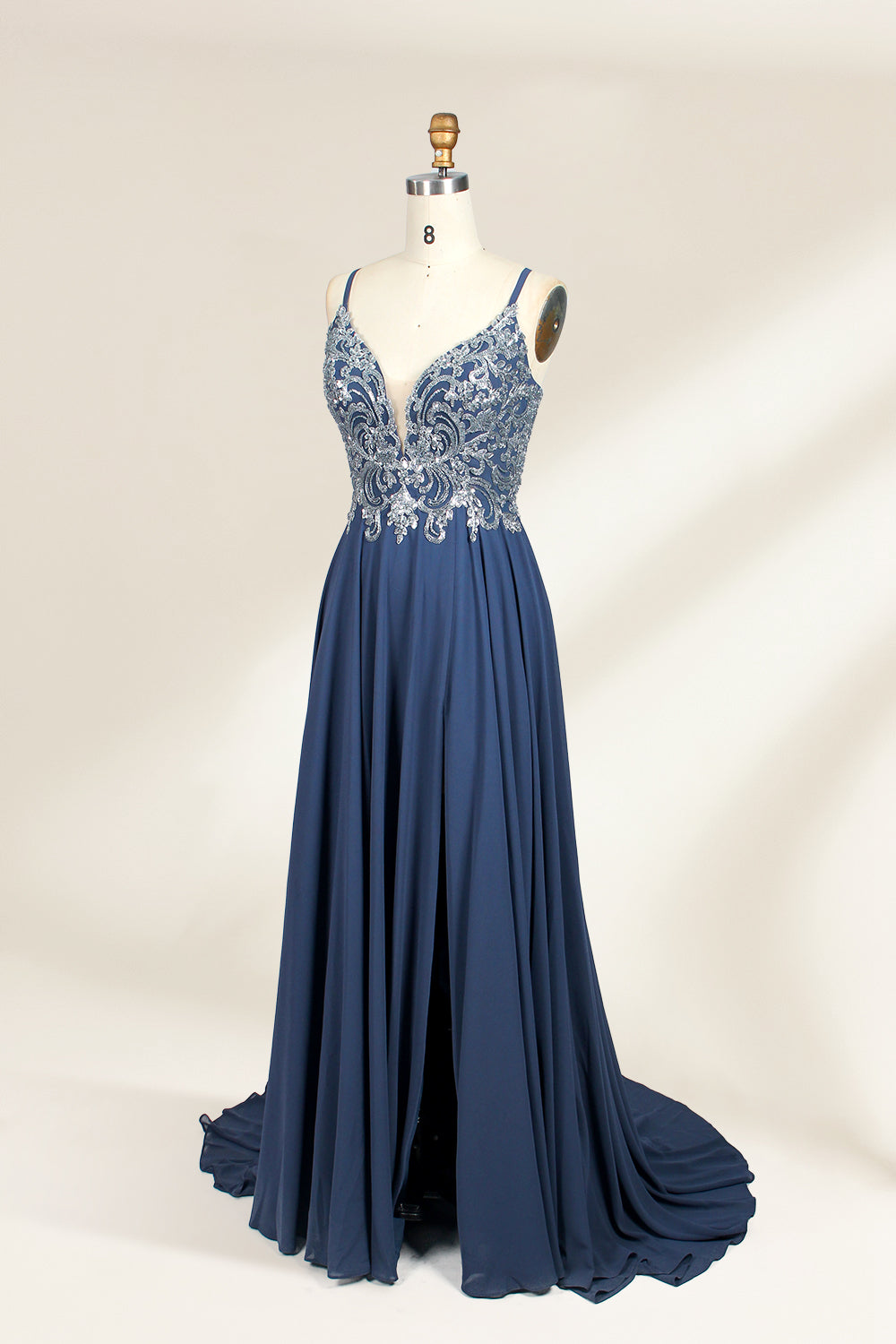 Photograph of a mannequin showcasing an elegant, floor-length evening gown. The gown features a deep V-neckline adorned with intricate blue floral embroidery, transitioning into a flowing, dark blue chiffon skirt. The mannequin stands against a plain, cream-colored backdrop. The dress exudes sophistication and luxury, perfect for formal events.