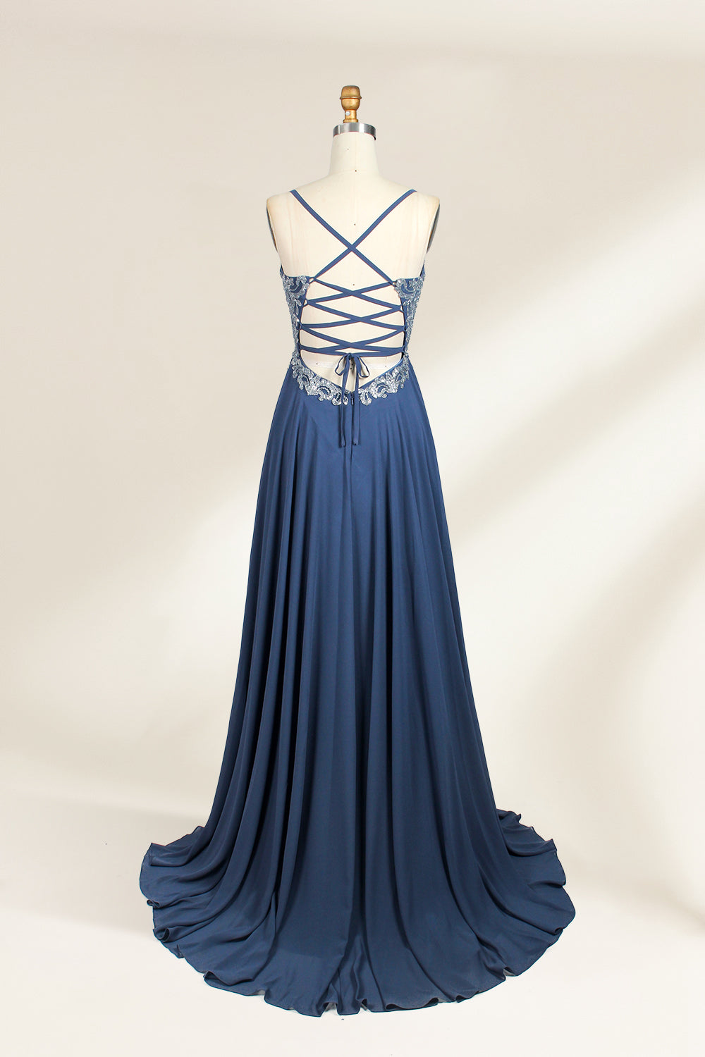 Photograph of a mannequin showcasing an elegant, floor-length evening gown. The gown features a deep V-neckline adorned with intricate blue floral embroidery, transitioning into a flowing, dark blue chiffon skirt. The mannequin stands against a plain, cream-colored backdrop. The dress exudes sophistication and luxury, perfect for formal events.