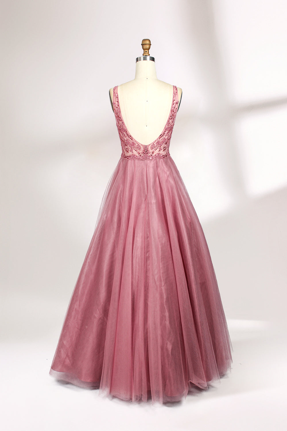 Photograph of a mannequin showcasing a floor-length, sleeveless, pink tulle gown with intricate lace detailing on the bodice and neckline. The gown features a fitted waist and flares out into a voluminous skirt, creating a ballerina-style silhouette. The mannequin stands against a plain white background with soft, diffused lighting casting subtle shadows.02