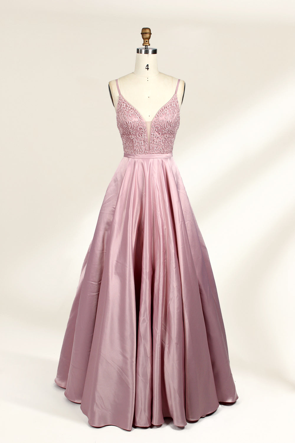Photograph of a stylish, sleeveless, floor-length satin gown on a dress form. The dress features a deep V-neckline adorned with intricate lace, and a high-waisted bodice that flares into a voluminous skirt. The gown is a soft, blush pink color. The background is a plain, light beige, enhancing the dress's elegant design.01