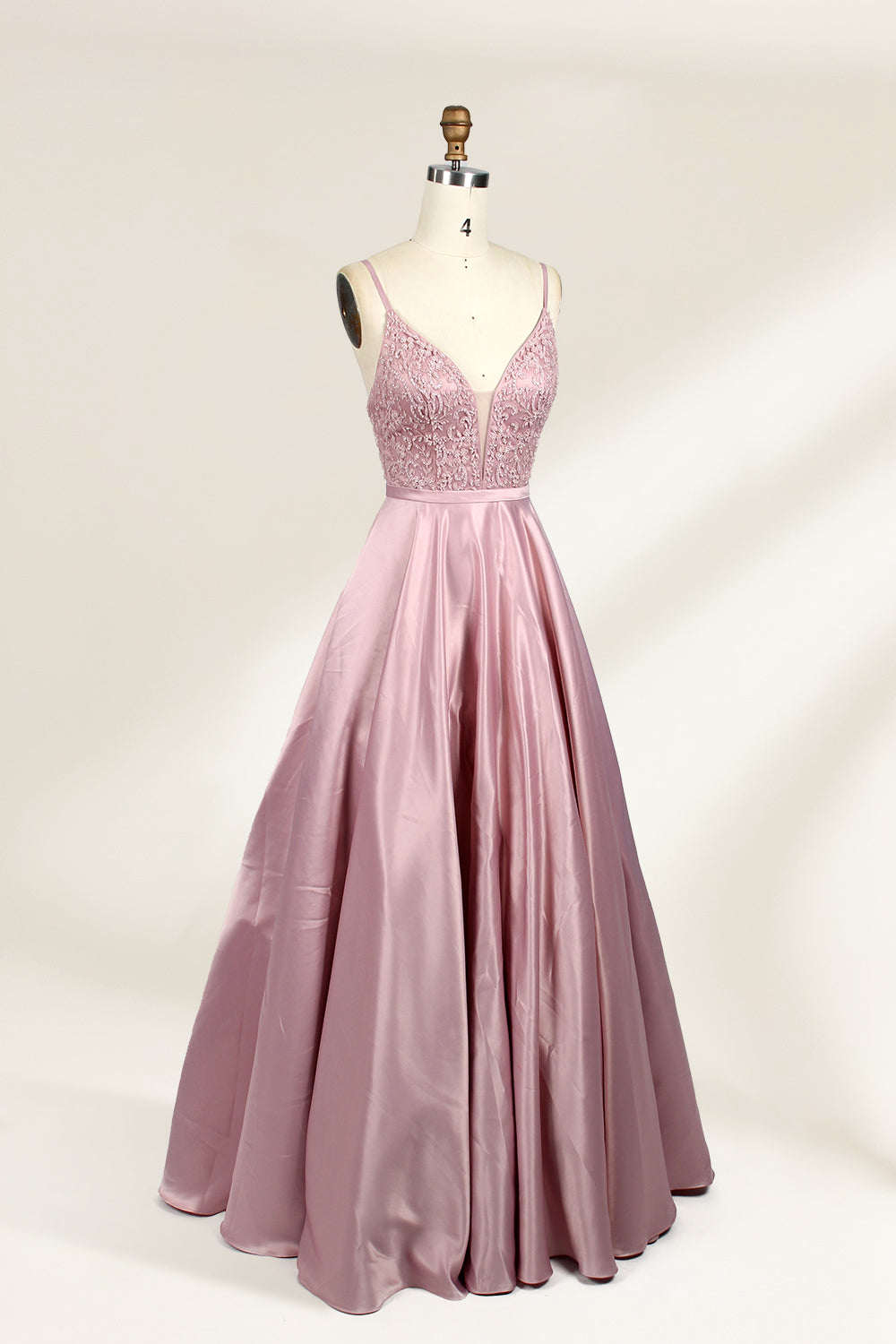 Photograph of a stylish, sleeveless, floor-length satin gown on a dress form. The dress features a deep V-neckline adorned with intricate lace, and a high-waisted bodice that flares into a voluminous skirt. The gown is a soft, blush pink color. The background is a plain, light beige, enhancing the dress's elegant design.02