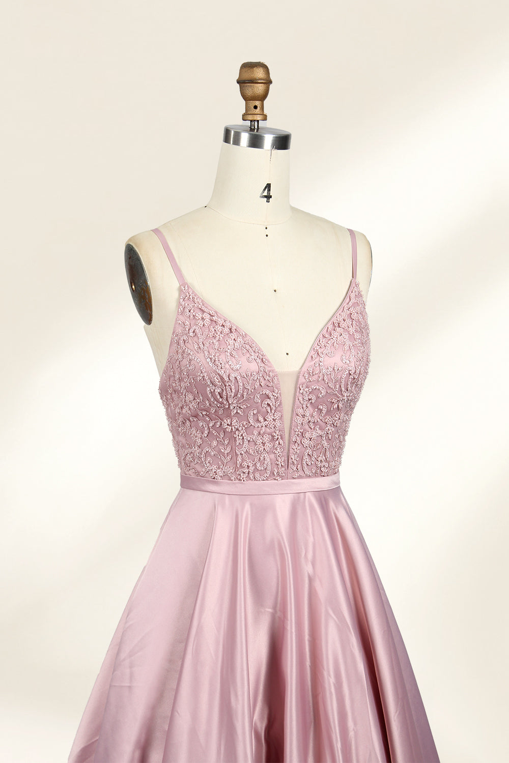 Photograph of a stylish, sleeveless, floor-length satin gown on a dress form. The dress features a deep V-neckline adorned with intricate lace, and a high-waisted bodice that flares into a voluminous skirt. The gown is a soft, blush pink color. The background is a plain, light beige, enhancing the dress's elegant design.023