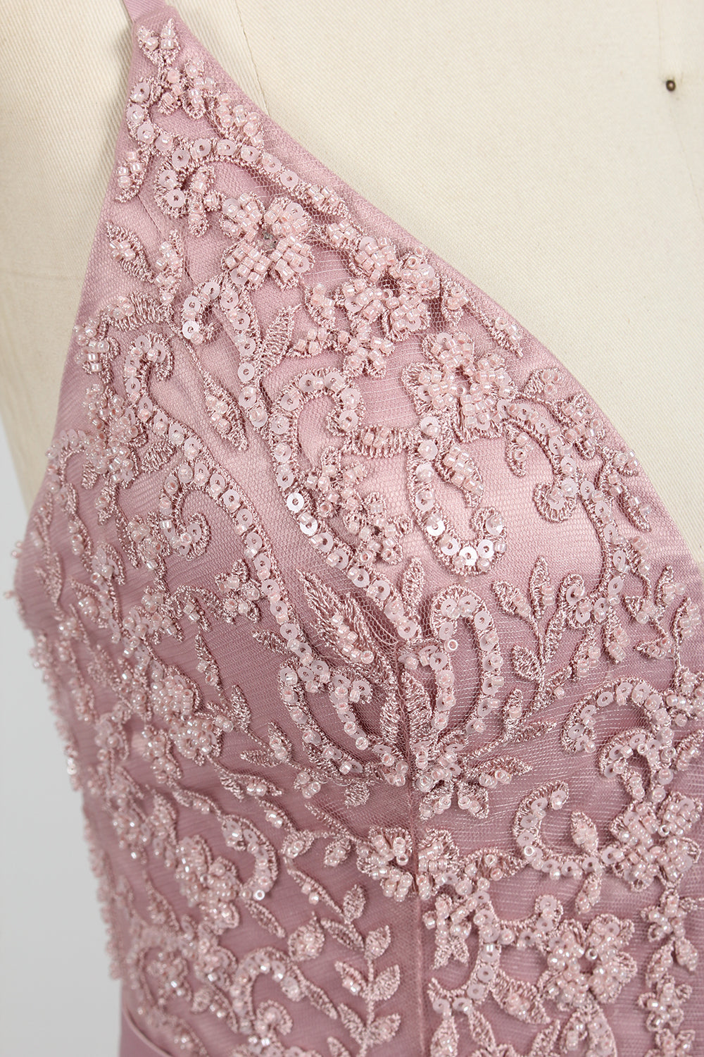 Photograph of a stylish, sleeveless, floor-length satin gown on a dress form. The dress features a deep V-neckline adorned with intricate lace, and a high-waisted bodice that flares into a voluminous skirt. The gown is a soft, blush pink color. The background is a plain, light beige, enhancing the dress's elegant design.04