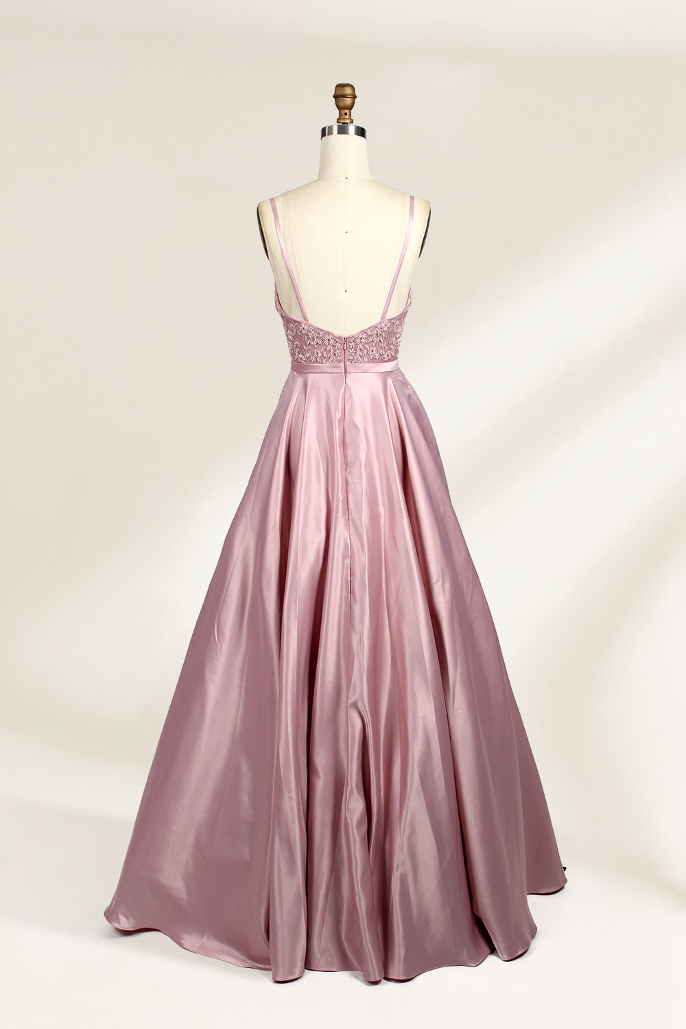 Photograph of a stylish, sleeveless, floor-length satin gown on a dress form. The dress features a deep V-neckline adorned with intricate lace, and a high-waisted bodice that flares into a voluminous skirt. The gown is a soft, blush pink color. The background is a plain, light beige, enhancing the dress's elegant design.05
