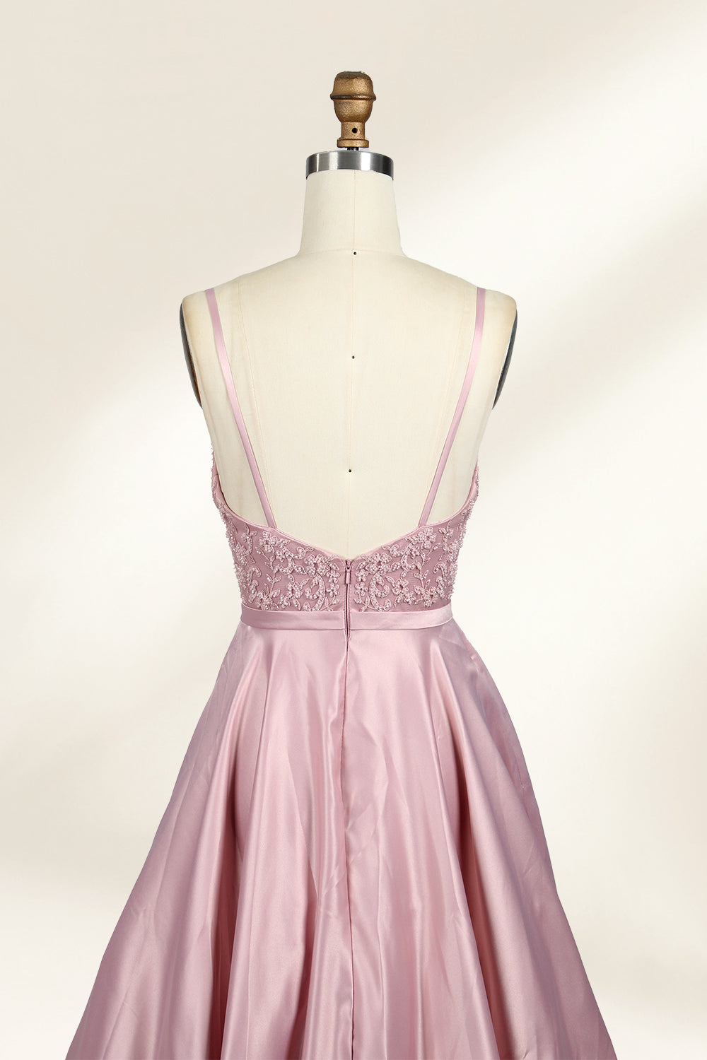 Photograph of a stylish, sleeveless, floor-length satin gown on a dress form. The dress features a deep V-neckline adorned with intricate lace, and a high-waisted bodice that flares into a voluminous skirt. The gown is a soft, blush pink color. The background is a plain, light beige, enhancing the dress's elegant design.06