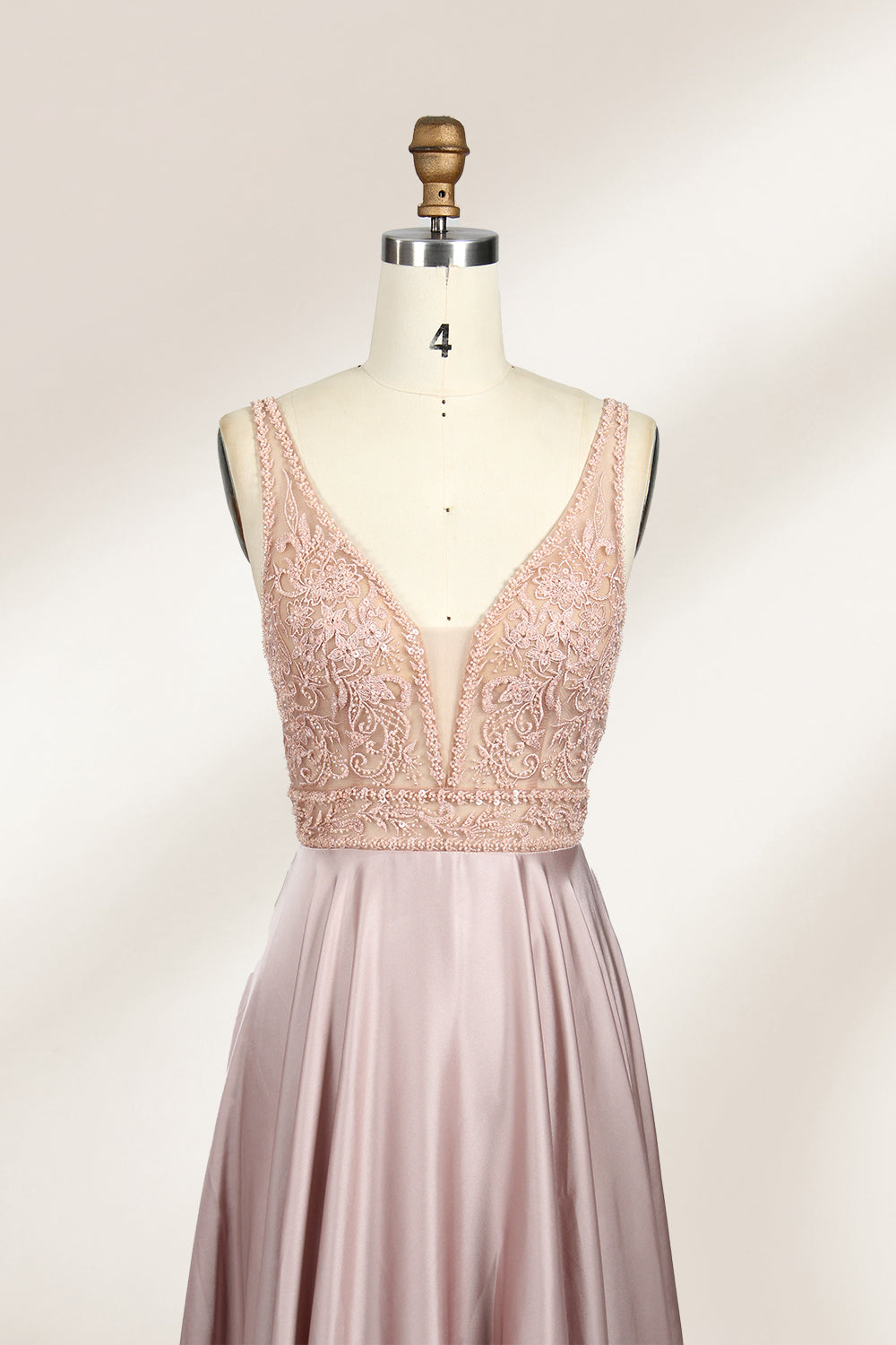 Photograph of a sleeveless, floor-length evening gown displayed on a dress form. The gown features a deep V-neckline adorned with intricate lace detailing and a sheer, blush pink fabric that flows into a smooth, satin skirt. The dress form is positioned against a plain, light beige background, emphasizing the gown's elegant design.03