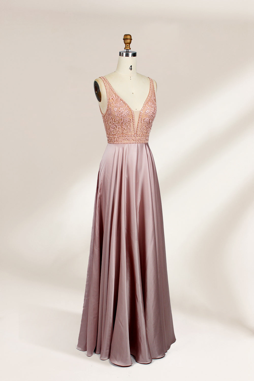 Photograph of a sleeveless, floor-length evening gown displayed on a dress form. The gown features a deep V-neckline adorned with intricate lace detailing and a sheer, blush pink fabric that flows into a smooth, satin skirt. The dress form is positioned against a plain, light beige background, emphasizing the gown's elegant design.04