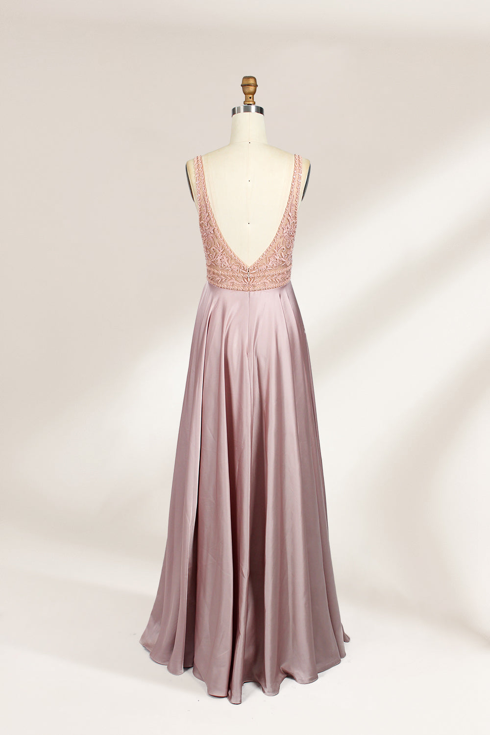 Photograph of a sleeveless, floor-length evening gown displayed on a dress form. The gown features a deep V-neckline adorned with intricate lace detailing and a sheer, blush pink fabric that flows into a smooth, satin skirt. The dress form is positioned against a plain, light beige background, emphasizing the gown's elegant design.02