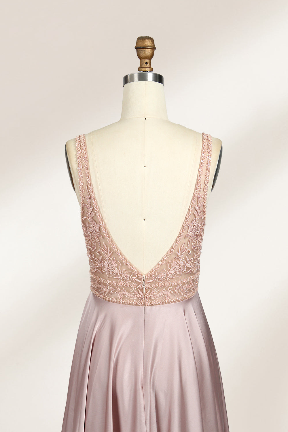 Photograph of a sleeveless, floor-length evening gown displayed on a dress form. The gown features a deep V-neckline adorned with intricate lace detailing and a sheer, blush pink fabric that flows into a smooth, satin skirt. The dress form is positioned against a plain, light beige background, emphasizing the gown's elegant design.06