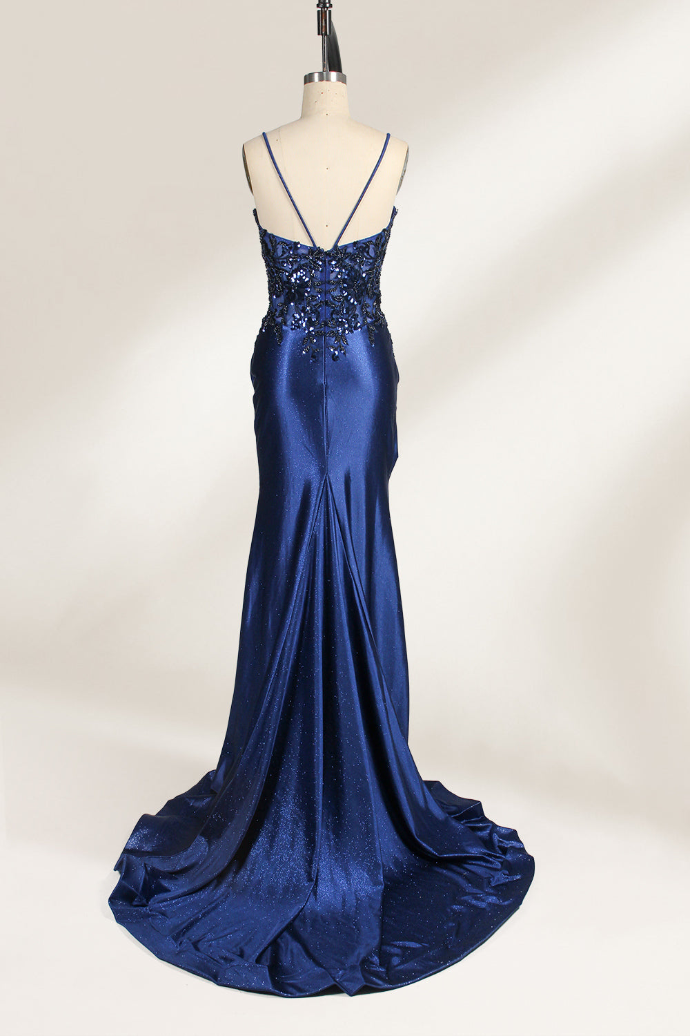 Photograph of a mannequin wearing a stunning, floor-length, navy blue satin evening gown. The gown features a deep V-back with thin straps and intricate sequin detailing along the back and neckline. The fitted bodice gradually flares into a flowing, elegant train. The mannequin stands against a plain, light beige backdrop, highlighting the gown's luxurious texture and shimmering finish. The gown exudes sophistication and glamour, ideal for formal events.2