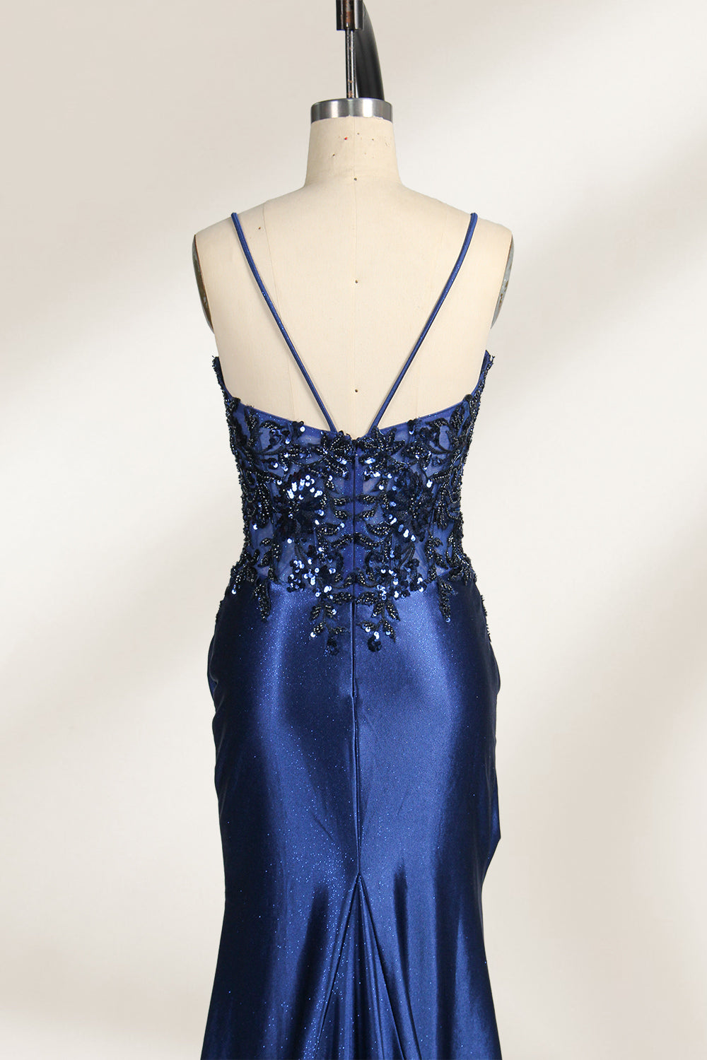Photograph of a mannequin wearing a stunning, floor-length, navy blue satin evening gown. The gown features a deep V-back with thin straps and intricate sequin detailing along the back and neckline. The fitted bodice gradually flares into a flowing, elegant train. The mannequin stands against a plain, light beige backdrop, highlighting the gown's luxurious texture and shimmering finish. The gown exudes sophistication and glamour, ideal for formal events.4